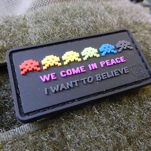 Könnte beinhalten: Schwarzer Patch mit einer Reihe von bunten, pixelartigen Space Invaders und dem Text "WE COME IN PEACE I WANT TO BELIEVE".