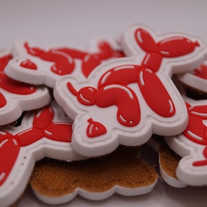 Könnte beinhalten: Eine Ansammlung roter Ballonhund-förmiger Aufnäher mit weißen Umrissen. Die Aufnäher sind aufeinander gestapelt und zeigen das rote Ballonhund-Design auf weißem Hintergrund. Die Aufnäher liegen auf einer braunen, strukturierten Oberfläche.