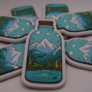 Könnte beinhalten: Eine Sammlung von teal- und weißfarbenen Aufnähern in Flaschenform, die jeweils ein Berglandschaftsdesign aufweisen. Der zentrale Aufnäher steht aufrecht, umgeben von anderen, die um ihn herum abgewinkelt sind. Das Design umfasst Berge, Bäume und Wasser.