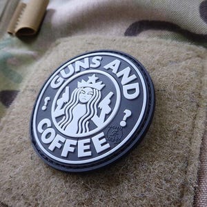Könnte beinhalten: Ein runder, schwarz-weißer Aufnäher mit den Worten "GUNS AND COFFEE" in einem stilisierten Schriftzug. Das Design enthält ein Logo, das an das eines Cafés erinnert, mit einer gekrönten Figur. Der Aufnäher ist auf einem Camouflage-Stoff befestigt.