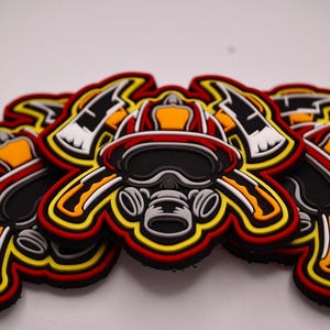 May include: A pile of firefighter-themed patches. Each patch features a firefighter helmet, axe, and respirator design with red, black, yellow, and white accents. The patches are layered on top of each other.