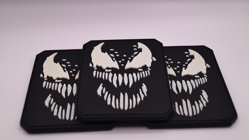 Peut inclure: Trois &eacute;cussons carr&eacute;s noirs avec un motif de visage Venom blanc. Les &eacute;cussons ont une apparence d&eacute;taill&eacute;e et textur&eacute;e, avec les &eacute;l&eacute;ments blancs se d&eacute;tachant sur le fond noir. Le design comprend des dents pointues et une expression mena&ccedil;ante.