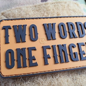 May include: A tan-colored patch with black text that reads "TWO WORDS ONE FINGER".