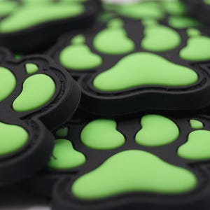 Puede incluir: Primer plano de varios parches negros con huellas de patas con almohadillas verdes brillantes. Los parches están hechos de un material gomoso y están dispersos sobre una superficie blanca. Los parches son probablemente para dueños de mascotas o amantes de los animales.