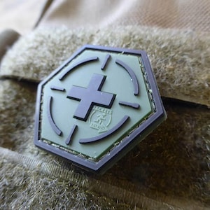 May include: A green and black hexagonal patch with a cross in the centre and the text "Jackets To Go" below. The patch has a black border and a black target design around the cross.