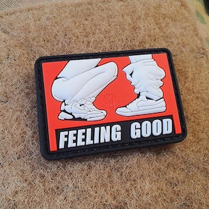 Op de afbeelding: Een rechthoekige patch met een zwarte rand en een rode achtergrond. De patch heeft een afbeelding van twee paar benen en voeten in witte sneakers. De woorden "FEELING GOOD" zijn in het wit gedrukt.