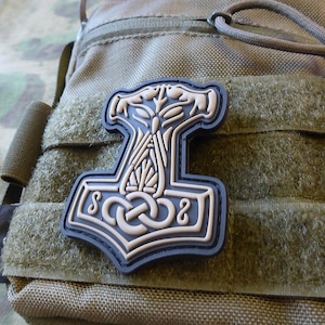 May include: A tan and black embroidered patch with a detailed design of a hammer, a knot, and a stylized tree. The patch is attached to a brown tactical bag.