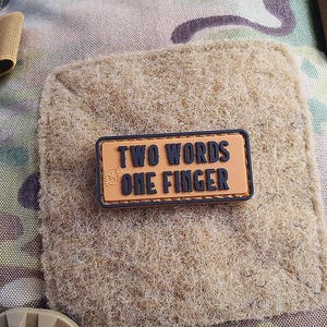 Könnte beinhalten: Ein braunes Patch mit einem schwarzen, gesteppten Rand und dem Text "TWO WORDS ONE FINGER" in schwarzen Buchstaben auf einem beigefarbenen Hintergrund.