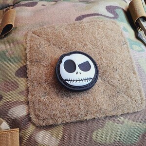 Puede incluir: Un parche de Jack Skellington en blanco y negro con una sonrisa cosida. El parche está unido a un parche de tela marrón con cierre de velcro.