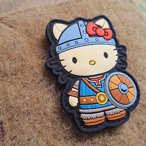 Può includere: Una toppa colorata raffigurante un personaggio di gatto dei cartoni animati vestito da guerriero vichingo. Il gatto indossa un elmo blu, un fiocco rosso e porta uno scudo. La toppa ha un bordo nero ed è ambientata su uno sfondo strutturato.