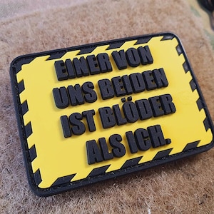 Pode incluir: Um patch retangular com fundo amarelo e texto preto que diz "EINER VON UNS BEIDEN IST BLÖDER ALS ICH." O patch tem uma borda preta com detalhes de chevron amarelos e pretos. O patch está sobre uma superfície texturizada bege.