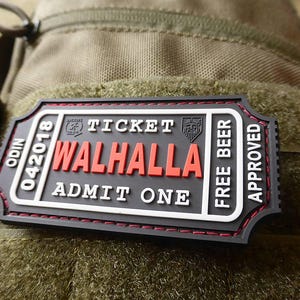 Könnte beinhalten: Schwarzes Ticket mit weißem Text, der "Odin 042018 Ticket Valhalla Admit One Free Beer Approved" lautet. Das Ticket ist an einem grünen Tarnstoff befestigt.