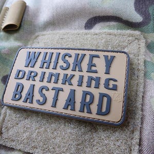 May include: A tan-coloured patch with black stitching and the text "Whiskey Drinking Bastard" in black lettering. The patch has a small logo in the bottom right corner.