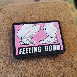 Op de afbeelding: Een rechthoekige patch met een roze achtergrond en de woorden "FEELING GOOD" in het wit. De patch toont een cartoonillustratie van twee paar benen en sneakers. De patch heeft een zwarte rand en ligt op een bruin, getextureerd oppervlak.