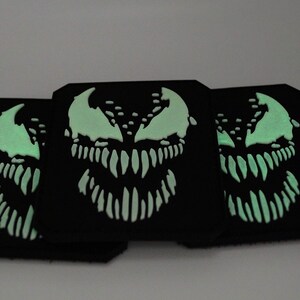 Peut inclure: Trois dessous de verre carr&eacute;s noirs avec un motif de visage Venom vert fluorescent. Le motif pr&eacute;sente un sourire mena&ccedil;ant et des dents ac&eacute;r&eacute;es. Les dessous de verre sont dispos&eacute;s en chevauchement, mettant en valeur les d&eacute;tails.