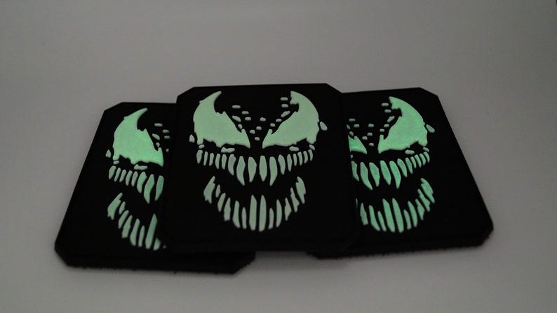 Peut inclure: Trois dessous de verre carr&eacute;s noirs avec un motif de visage Venom vert fluorescent. Le motif pr&eacute;sente un sourire mena&ccedil;ant et des dents ac&eacute;r&eacute;es. Les dessous de verre sont dispos&eacute;s en chevauchement, mettant en valeur les d&eacute;tails.