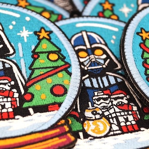 Könnte beinhalten: Ein Set mit vier gestickten Weihnachts-Patches mit Darth Vader und anderen Star Wars-Charakteren. Die Patches sind rund und haben ein Schneekugel-Design mit blauem Hintergrund und weißem Schnee. Jeder Patch zeigt einen anderen Charakter und einen Weihnachtsbaum.