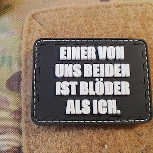 Könnte beinhalten: Ein schwarzer rechteckiger Aufnäher mit weißem Text "EINER VON UNS BEIDEN IST BLÖDER ALS ICH." Der Aufnäher hat einen genähten Rand und befindet sich vor einem Tarn- und beigefarbenen Hintergrund.