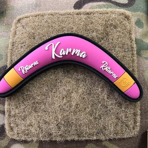 May include: Pink boomerang with the text "Karma Returns" on both sides. The boomerang is made of a soft, flexible material and has a yellow stripe on each side.