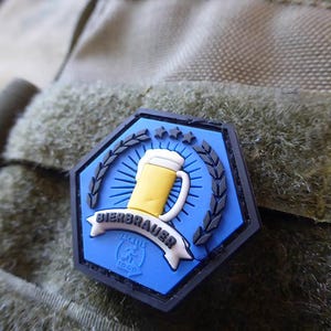 May include: Blue and black hexagonal patch with a white banner that says "Bierbrauer 24/7 To Go". The patch features a yellow beer mug with a white foam top and a black outline. The beer mug is surrounded by a black wreath with five white stars.