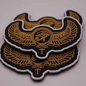 Könnte beinhalten: Ein Stapel schwarzer und goldener Emblem-Patches mit weißem Rand. Die Patches zeigen ein geflügeltes Design mit einem Augensymbol in der Mitte und einem Kreuz unten. Die Flügel und das Auge sind goldfarben.