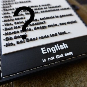 Pode incluir: Patch preto e branco com o texto "English is not that easy" e uma lista de frases em inglês. O patch tem uma borda costurada.