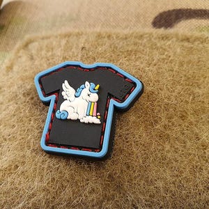 Könnte beinhalten: Ein schwarzer T-Shirt-Patch mit blauem Rand, der ein weißes Einhorn mit Regenbogen-Erbrochenem zeigt. Der Patch hat rote Nähte an den Rändern.