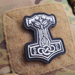 Könnte beinhalten: Ein weiß-schwarzer Mjolnir-Patch, der Hammer von Thor. Der Patch ist aus einem gummiartigen Material gefertigt und hat ein detailliertes Design. Der Patch ist auf einem braunen Stoffhintergrund befestigt.