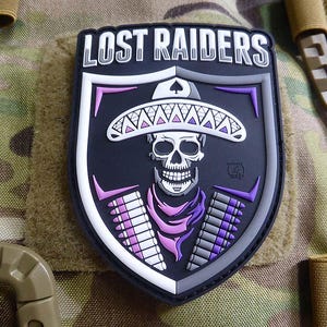 May include: A black and white patch with the text "LOST RAIDERS" in white. The patch features a skull wearing a sombrero with purple, pink, and white accents. The skull is surrounded by a purple and white bandolier.
