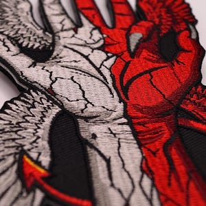 Könnte beinhalten: Gestickter Aufnäher mit geteiltem Design: ein weißer Engelsflügel und eine rote Teufelshand. Der Engelsflügel hat eine rissige Textur, während die Teufelshand eine gekrallte Hand und einen roten Schwanz hat. Der Aufnäher ist schwarz umrandet.
