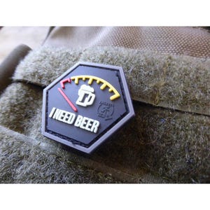 May include: A hexagonal patch with the words "I NEED BEER" in white lettering. The patch has a beer mug icon and a gauge design, with yellow and red accents. It is attached to a textured, olive-green fabric.