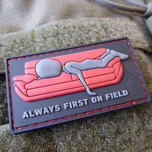 May include: A black and red embroidered patch with a cartoon figure lying on a red couch. The text "ALWAYS FIRST ON FIELD" is printed on the patch.
