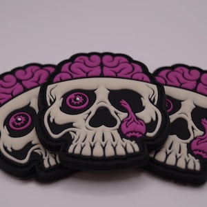 May include: Three skull-shaped patches with a black border, off-white skull, and a purple brain design. The eye sockets and tear ducts are also purple. The patches are stacked on top of each other.