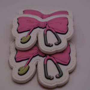 May include: Several pink bow-shaped shoe charms with white outlines and gray and green accents are stacked on top of each other. The charms have a detailed design, with the bow's ribbon and dangling elements clearly visible.