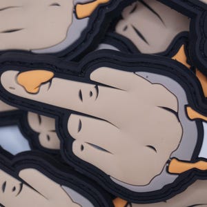 Könnte beinhalten: Nahaufnahme mehrerer Aufnäher mit einer Hand, die den Mittelfinger zeigt. Die Aufnäher haben eine beige Hand mit schwarzer Umrandung und schwarzem Rand. Der Fingernagel ist orange.