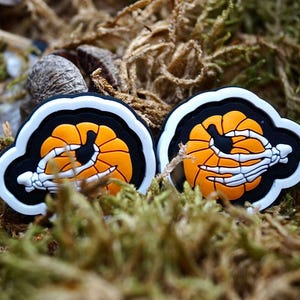 Könnte beinhalten: Zwei weiße und schwarze Halloween-Anhänger mit einer Skeletthand, die einen Kürbis hält. Der Kürbis ist orange mit einem schwarzen Stiel.