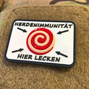 Peut inclure: Un patch blanc avec du texte noir et un motif en spirale rouge et blanc. Le texte dit "HERDENIMMUNITÄT HIER LECKEN".