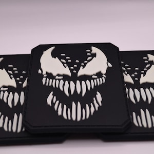 Peut inclure: Trois écussons rectangulaires noirs présentant un motif blanc en relief représentant une créature souriante avec des dents pointues et de grands yeux. Les écussons ont des coins arrondis et une finition mate, affichant un design contrasté.