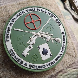 Könnte beinhalten: Ein runder grüner und weißer Patch mit einem Scharfschützengewehr und einer Spielkarte mit dem Ass der Pik. Der Text auf dem Patch lautet "From a place you will not see comes a sound you will not hear".