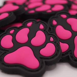 Könnte beinhalten: Nahaufnahme mehrerer schwarzer und pinkfarbener Pfotenabdruck-Patches. Die Patches haben einen schwarzen Rand und ein erhabenes pinkfarbenes Pfotenabdruck-Design. Die Patches sind auf einer weißen Oberfläche verstreut.