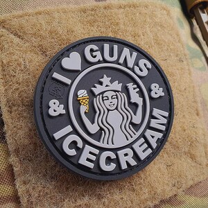 Op de afbeelding: Een zwarte en grijze patch met de tekst "I ♥ GUNS & ICE CREAM" in witte letters. De patch toont een gestileerde afbeelding van een zeemeermin met een kroon en een pistool, die een ijshoorn vasthoudt.