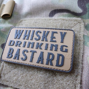 May include: A tan-coloured patch with black stitching and the text "Whiskey Drinking Bastard" in black lettering. The patch has a small logo in the bottom right corner.