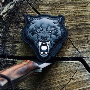 May include: A black and gray wolf patch with an open mouth and visible teeth. The patch is placed on a weathered wooden surface, with a knife in the foreground.