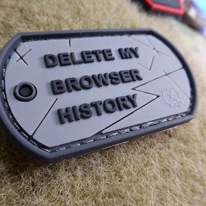 Könnte beinhalten: Ein grau-schwarzer Aufnäher in Form einer Erkennungsmarke mit dem Text "DELETE MY BROWSER HISTORY" in fetten, schwarzen Buchstaben. Der Aufnäher hat einen schwarzen Rand und ein kleines kreisförmiges Detail auf der linken Seite. Der Hintergrund ist ein strukturiertes, beiges Gewebe.