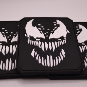 Peut inclure: Trois écussons rectangulaires noirs avec un motif de visage Venom blanc. Le motif présente un visage blanc avec des dents pointues et une expression menaçante. Les écussons sont empilés les uns sur les autres.