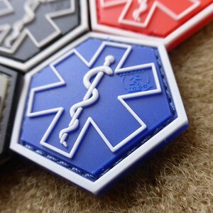 Könnte beinhalten: Blau-weißer Lebensstern-Patch mit einem weißen medizinischen Caduceus-Symbol in der Mitte. Der Patch hat einen weißen Rand und ist auf einen braunen Stoff genäht.