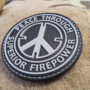 Könnte beinhalten: Ein schwarz-weißes Friedenssymbol-Patch mit dem Text "Peace Through Superior Firepower" um den Rand herum. Das Patch hat einen genähten Rand.
