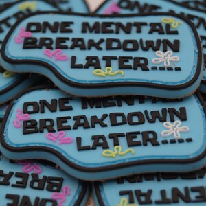 Könnte beinhalten: Mehrere hellblaue Kekse mit dem Text "ONE MENTAL BREAKDOWN LATER..." in Schwarz. Jeder Keks hat einen schwarzen Rand und ist mit rosa, weißen und gelben Blumenmustern verziert.