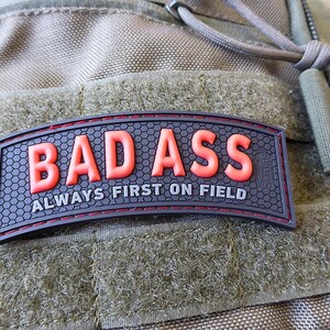 Könnte beinhalten: Ein schwarzer und roter Aufnäher mit den Worten "BAD ASS" in fetten, orangefarbenen Buchstaben. Darunter steht der Satz "ALWAYS FIRST ON FIELD" in Weiß. Der Aufnäher befindet sich auf einem grün-beigen strukturierten Hintergrund.