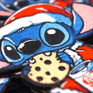 Puede incluir: Un parche bordado azul y rojo de un personaje de dibujos animados que lleva un gorro de Papá Noel y sostiene una galleta. El personaje tiene ojos grandes y una gran sonrisa. El parche está hecho de fieltro y tiene un borde cosido.